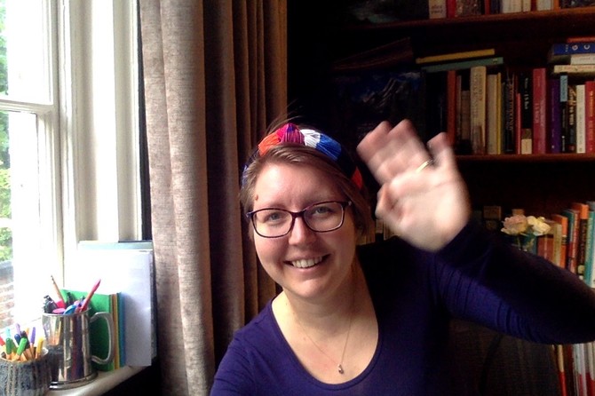 Natasha wears a colourful headscarf, dark-rimmed glasses and a blue long-sleeved top, and is smiling and waving at the camera