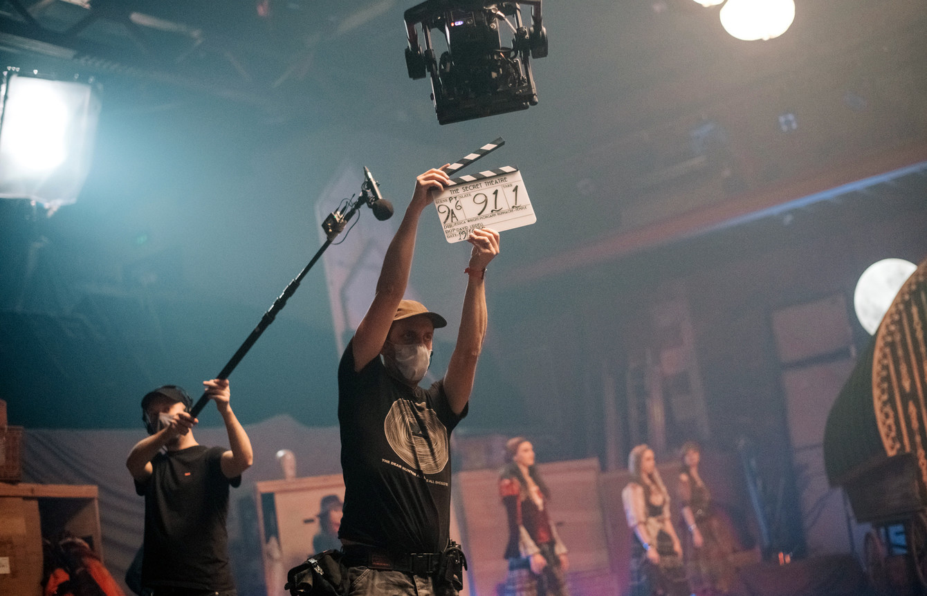 A film crew member holds a film slate aloft, while a boom operator stands behind them, and costumed cast members are poised in the background