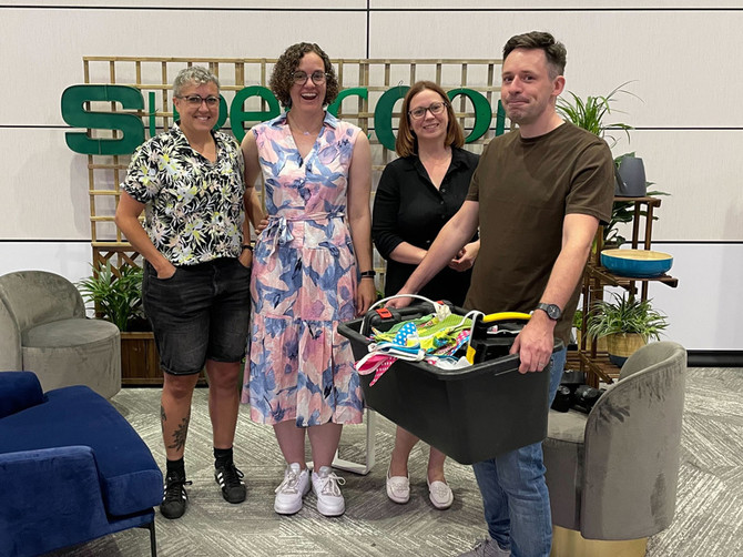 Left to right are a smiling Hayley, Kate, and Ceri. Then James who has a confused look on his face. They're standing in front of garden trellis with big green letters, spelling 'Supercool', attached to it.