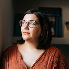 A white woman with black, shoulder-length hair and glasses. She's wearing a deep orange top and is turned to look a little to the left – towards a light source.