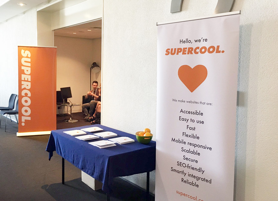 Supercool Ama Conf 2019 Consultancy Pod View from outside the Supercool Pod at AMAConf 2019. There're a couple of Supercool pull-up banners, a table with our 'Access Notebooks' and some oranges, and James is sat inside the pod running a session.
